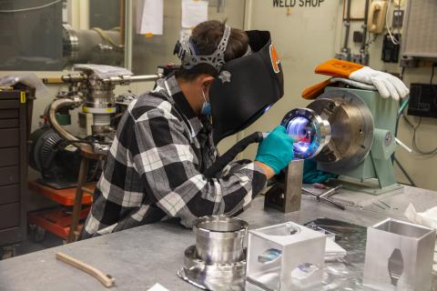 Image of a person welding, a blue glow emanates from the round piece of metal being welded