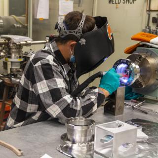 Image of a person welding, a blue glow emanates from the round piece of metal being welded